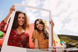 Two smiling women standing together and holding empty frame outdoors 0yNmj5