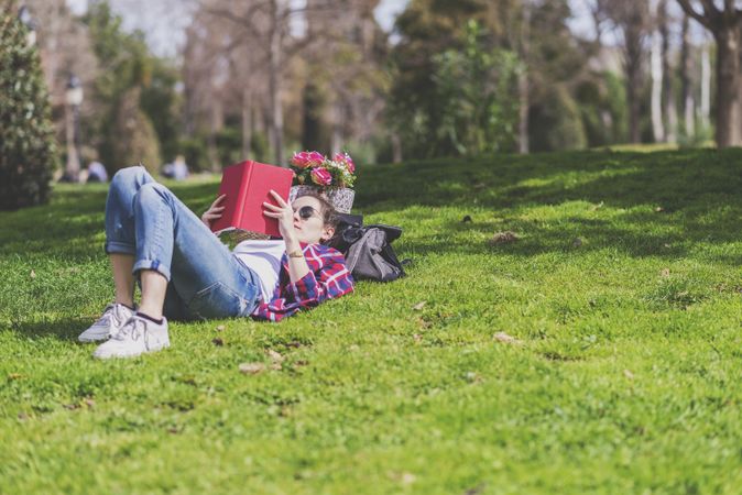 Side view of a hipster happy woman lying on grass in sunny day at park while reading a red book