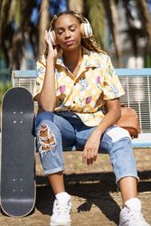 Female in bold patterned shirt sitting on park bench listening to music on large headphones