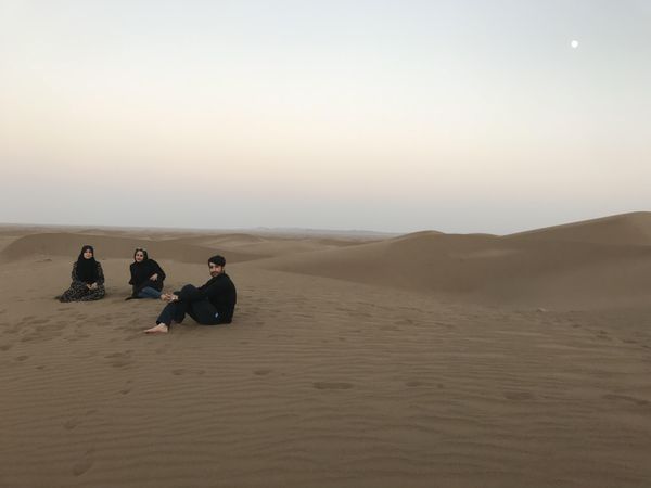 Two women wearing hijab and a man sitting on sand in desert