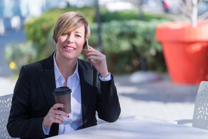 Smiling female talking on phone sitting at table outside with coffee