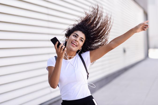 Arab woman in sport clothes with curly hair walking outside while dancing next to wall
