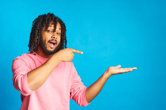 young man with braided hair happy with surprised gesture pointing isolated on blue background