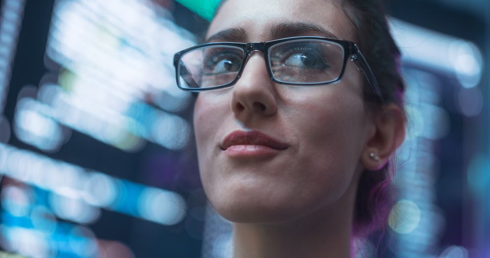 Close Up Portrait of Smiling Woman Looking Around, Lines of Code Language Reflected on her Glasses from Big Display Screens Surrounding her.