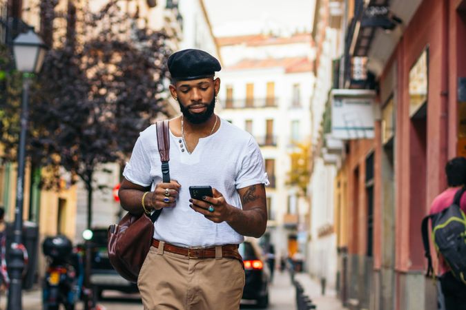 Stylish Black man walking on the city street using smartphone
