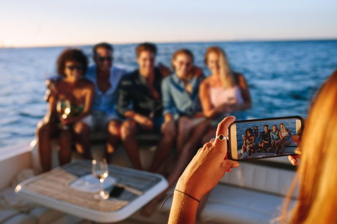 Close up of woman holding smart phone and taking picture of friends sitting on boat