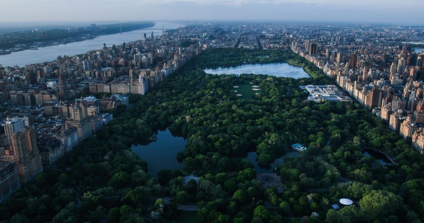 Aerial Helicopter Photo Over Central Park with Nature, Trees, People Having Picnic and Resting on a Field Around Manhattan Skyscrapers Cityscape. Beautiful Evening with Warm Sunset Light