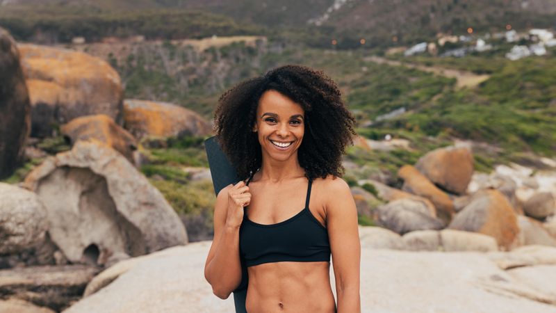 Fit happy Black woman standing in nature holding yoga mat