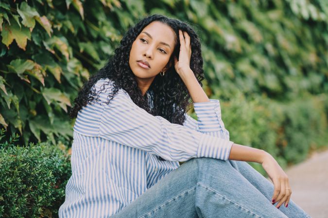 A contemplative young woman stands against a lush green ivy wall, surrounded by nature