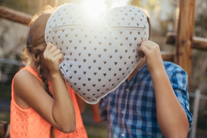 Close up of a boy and girl holding a heart shaped pillow hiding their heads behind it