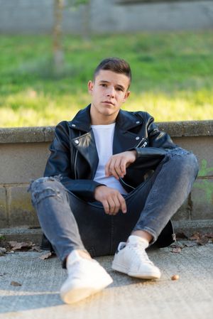 Teenage male wearing a leather jacket leaning on ledge outside looking at camera