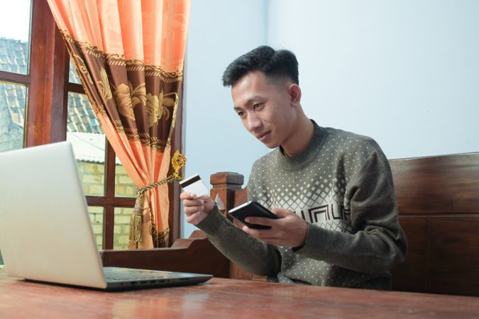 Serious Asian man in sweater shopping online holding credit card and smartphone