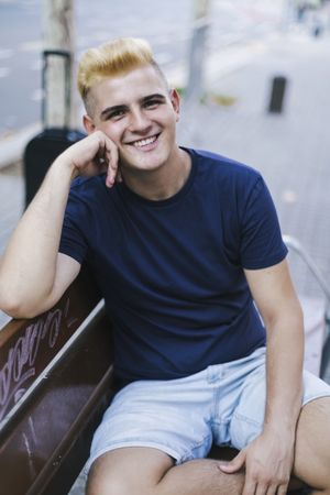 Portrait of a smiling young blonde male sitting on a bench in the street