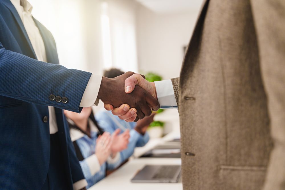 Close-up of two businessmen shaking hands in an office setting with applauding colleagues in the background, faces not visible