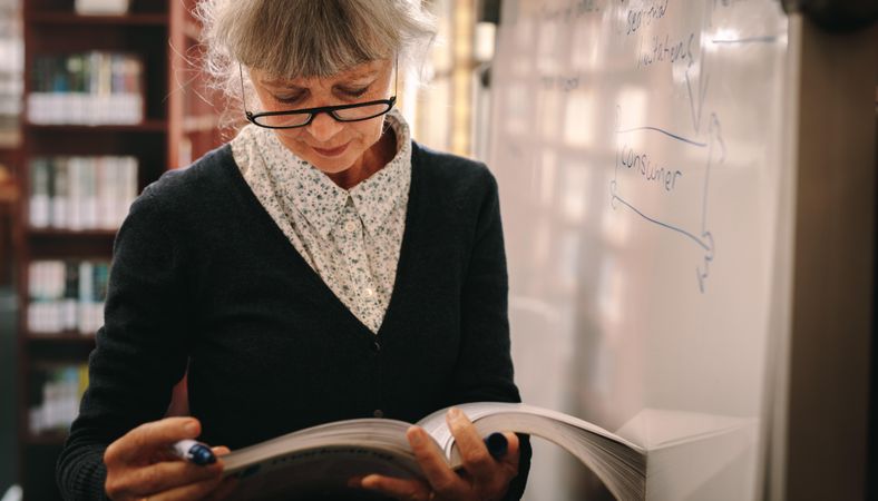 Close up of a mature woman standing near a whiteboard flipping pages of a text book