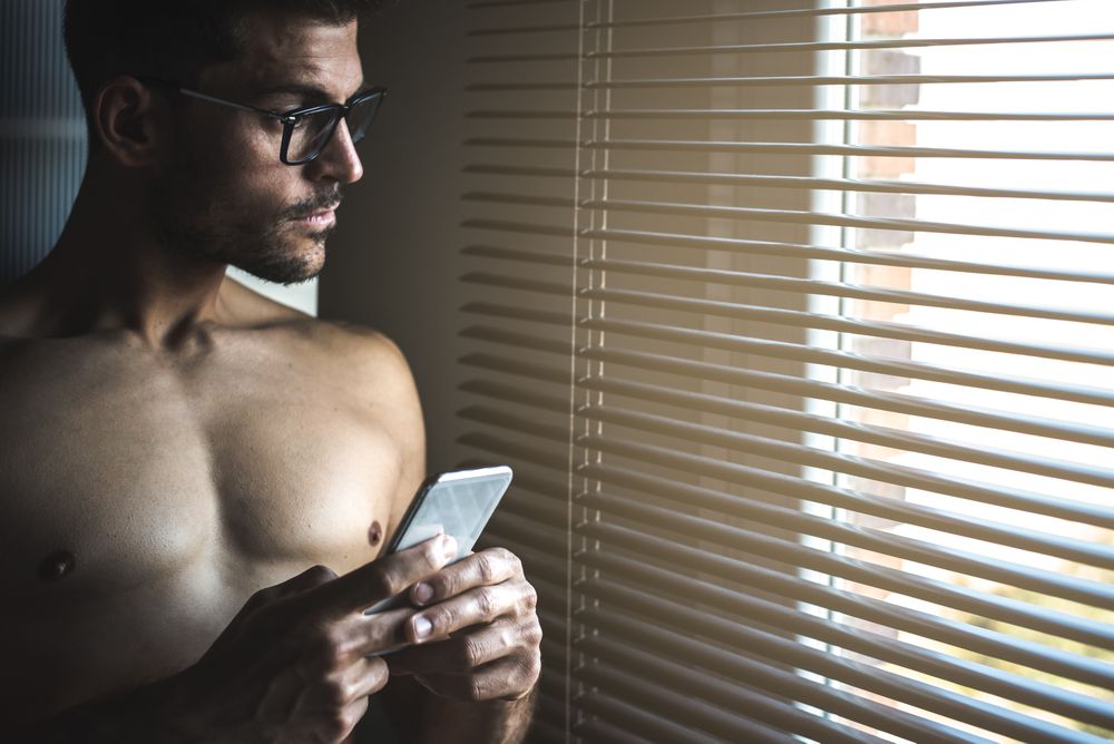 Man standing by a windows blind on the mobile phone