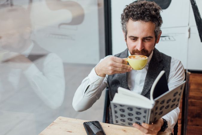 Man with coffee in cafe while reading book with smart phone