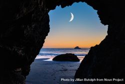 Crescent moon seen between rocky arch on quiet beach at dusk 5wEaRb