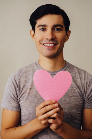 Romantic happy Hispanic male holding cut out pink heart to his chest, vertical