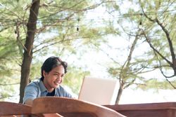 Happy Asian Young businessman work at outdoor with laptop. Concept of remote and freelance work. Young successful man wearing casual blue cloth.