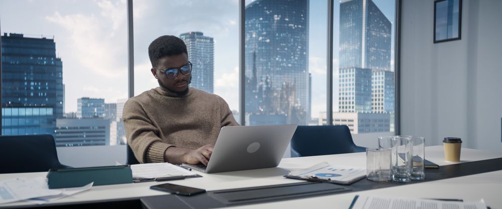 Portrait of Successful Black Businessman Working on Laptop Computer in His Big City Office.