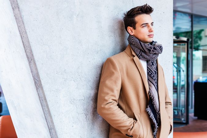 Serious man in camel coat and scarf looking away leaning on wall outside