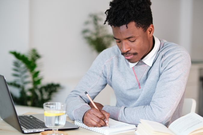 Black student writing in notebook near laptop at home