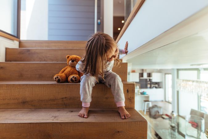 Cute little girl sitting on staircase and looking down through a glass wall indoors