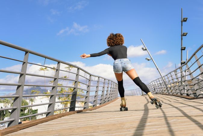 Back of woman with afro hairstyle in roller skates on wooden bridge