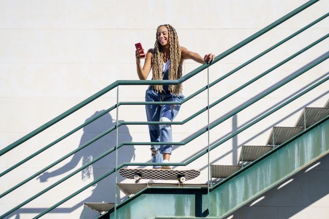 Smiling female skater standing and using phone on stairs, copy space