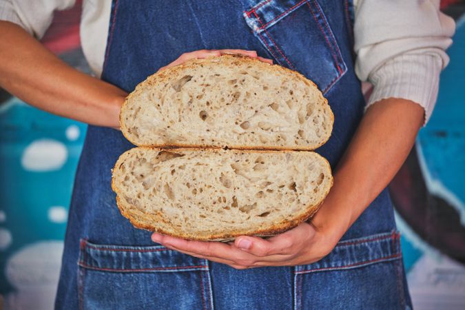 Photo detail of female hands holding an artisan bread broken in half with a blue kitchen apron.