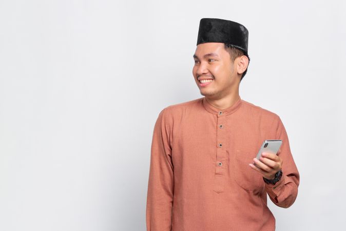 Muslim man in kufi hat holding mobile phone and looking away