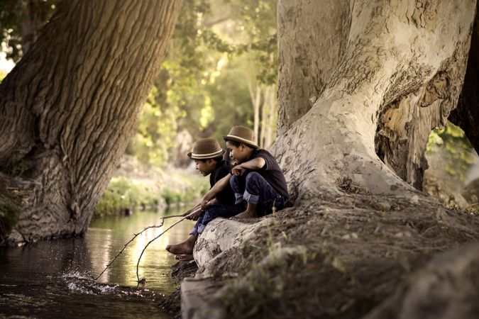 Two boys wearing brown hat sitting on rock near river