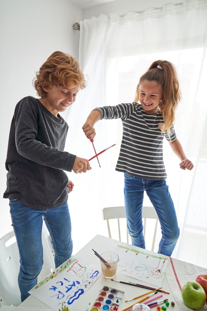 Happy children playing with paintbrushes near white table