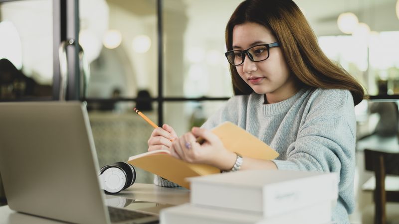 Asian female student in glasses looking at laptop and taking notes in a notebook in hand
