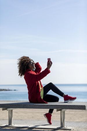 Side view of female taking picture on smart phone on bench on the coast, vertical composition