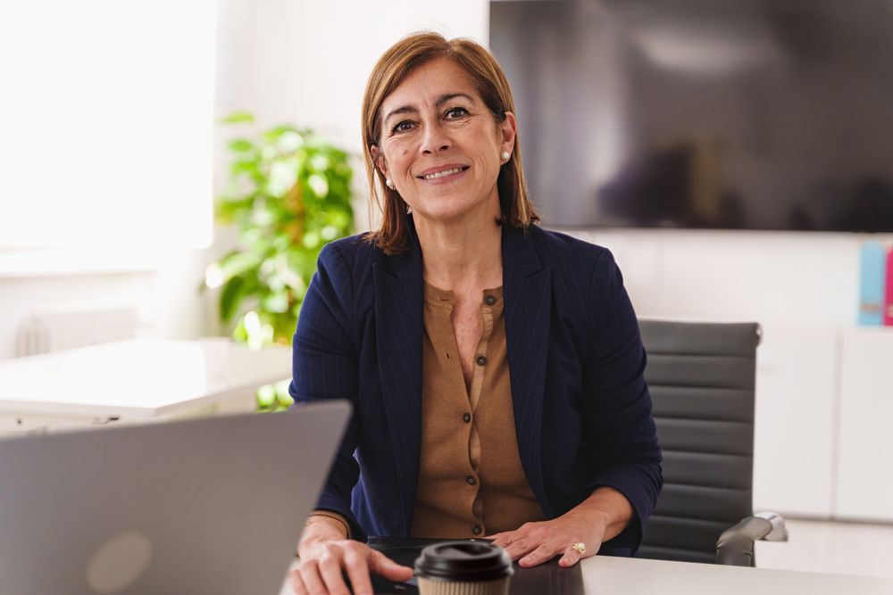 A woman in her 50s sits at a desk with a laptop, displaying a warm smile