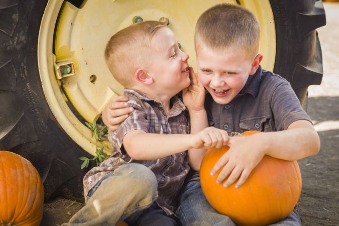 Two Boys Sitting Against Tractor Tire Holding Pumpkins Whispering Secrets