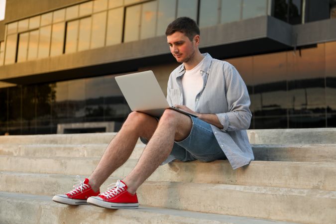 Young man sits in steps and working in laptop outdoor. Freelancer