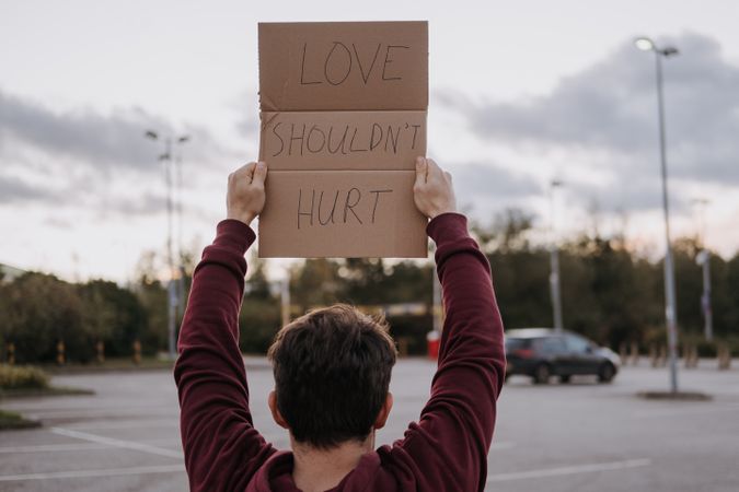 Back view of man holding cardboard paper with "Love Shouldn't Hurt" written on it