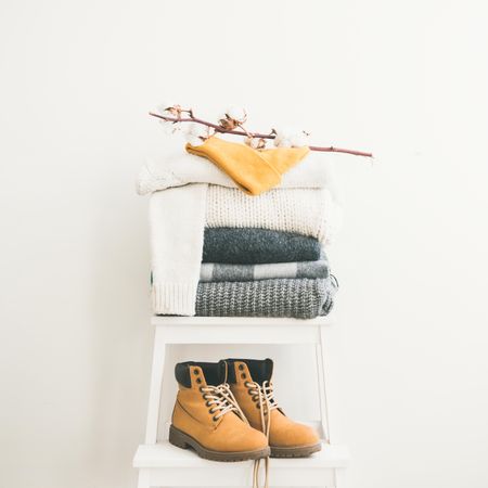 Pile of clean, folded sweaters on light background with yellow boots, dried cotton, square crop