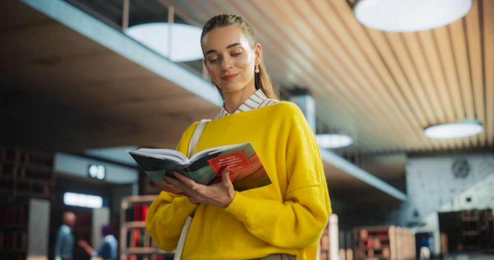 Young Happy Female Student Borrowed a Book From a College Library. Attractive Woman Hungry for Knowledge, Browsing the Pages, Reading Intellectual Texts and Essays by a Social Studies Professor