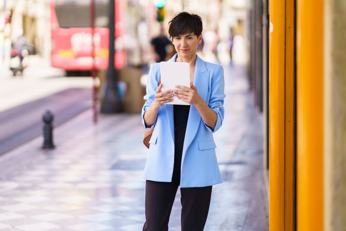 Woman in glasses and blue blazer standing on street looking at digital tablet