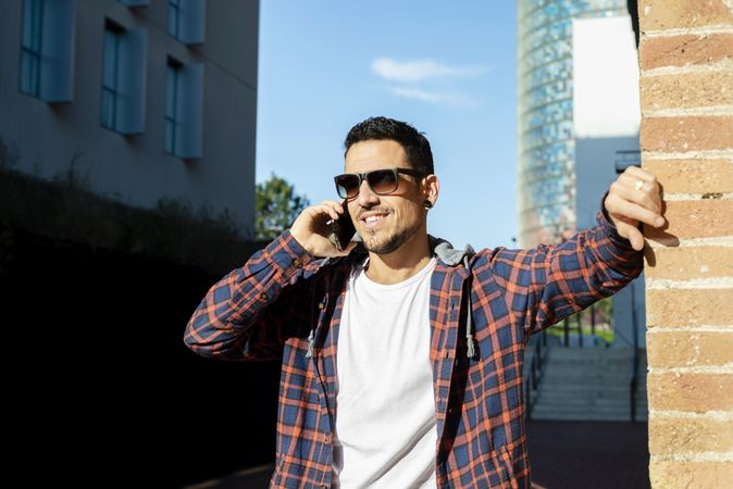 Man standing by brick wall wearing sunglasses and chatting on smartphone outdoors in the sun