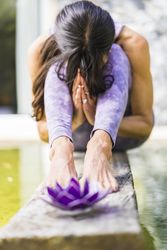 Aesthetic shot of a praying woman sitting behind a purple glass lotus on a tree trunk over the water.
