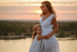 Woman and child in dresses hugging in front of lake during beautiful sunset
