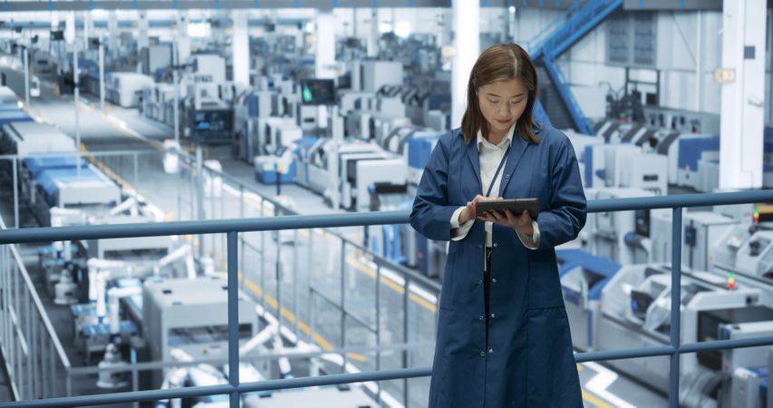 Japanese Factory Manager Working on a Tablet at a Futuristic Industrial Manufacture with Autonomous AI Robots. Specialist Monitoring All the Data on a Device, Checking Reports and Planning Research