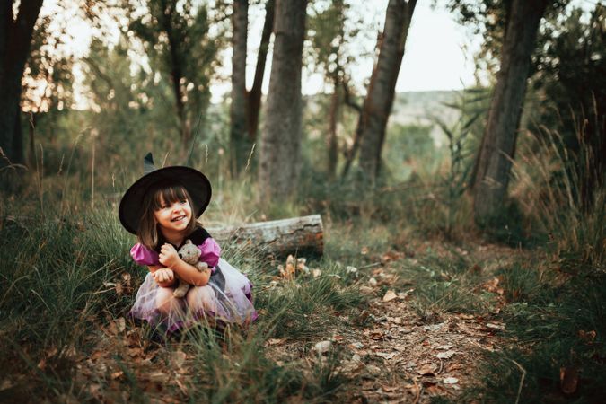 Laughing girl in witch costume crouched down in the forest with her teddy bear