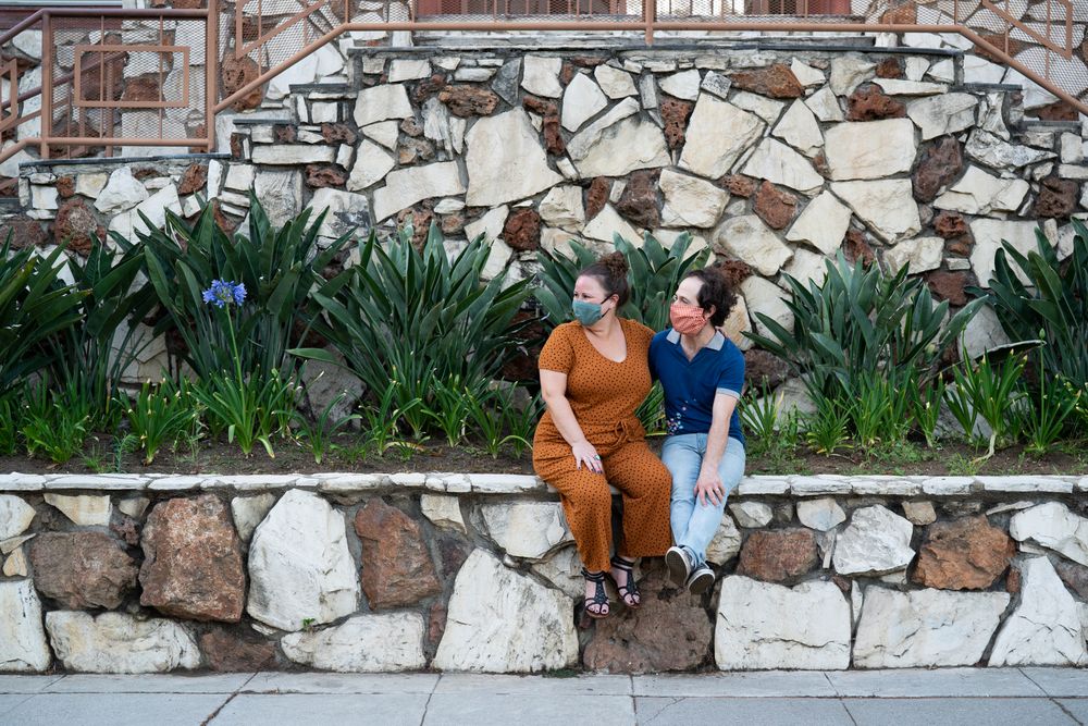 Happy couple sitting near sidewalk wearing masks and looking away