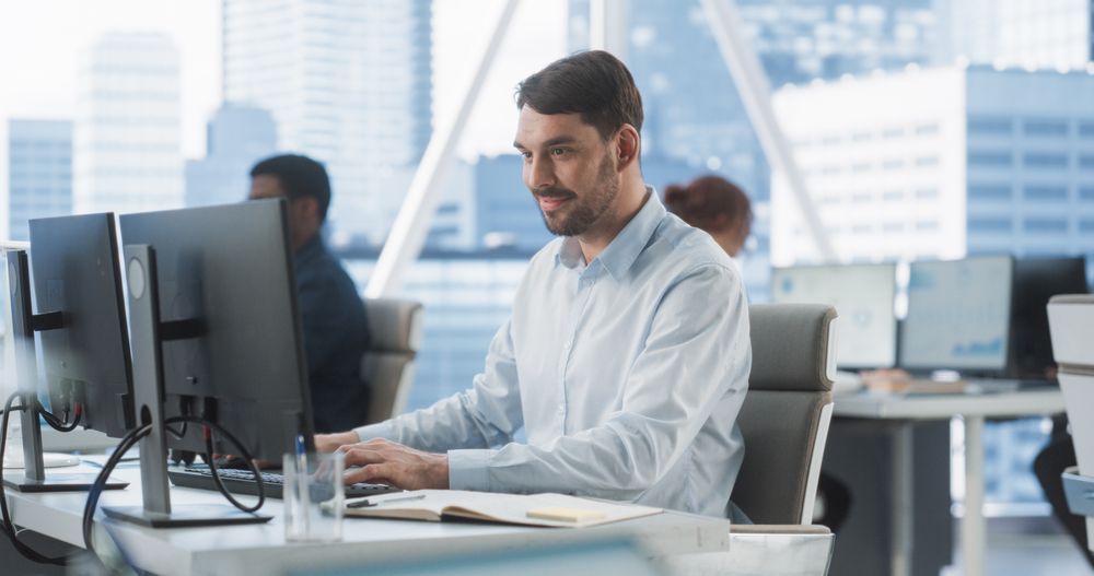 Happy Male Sales Manager Typing On Desktop Computer In Diverse Corporate Office With Megapolis Window View.